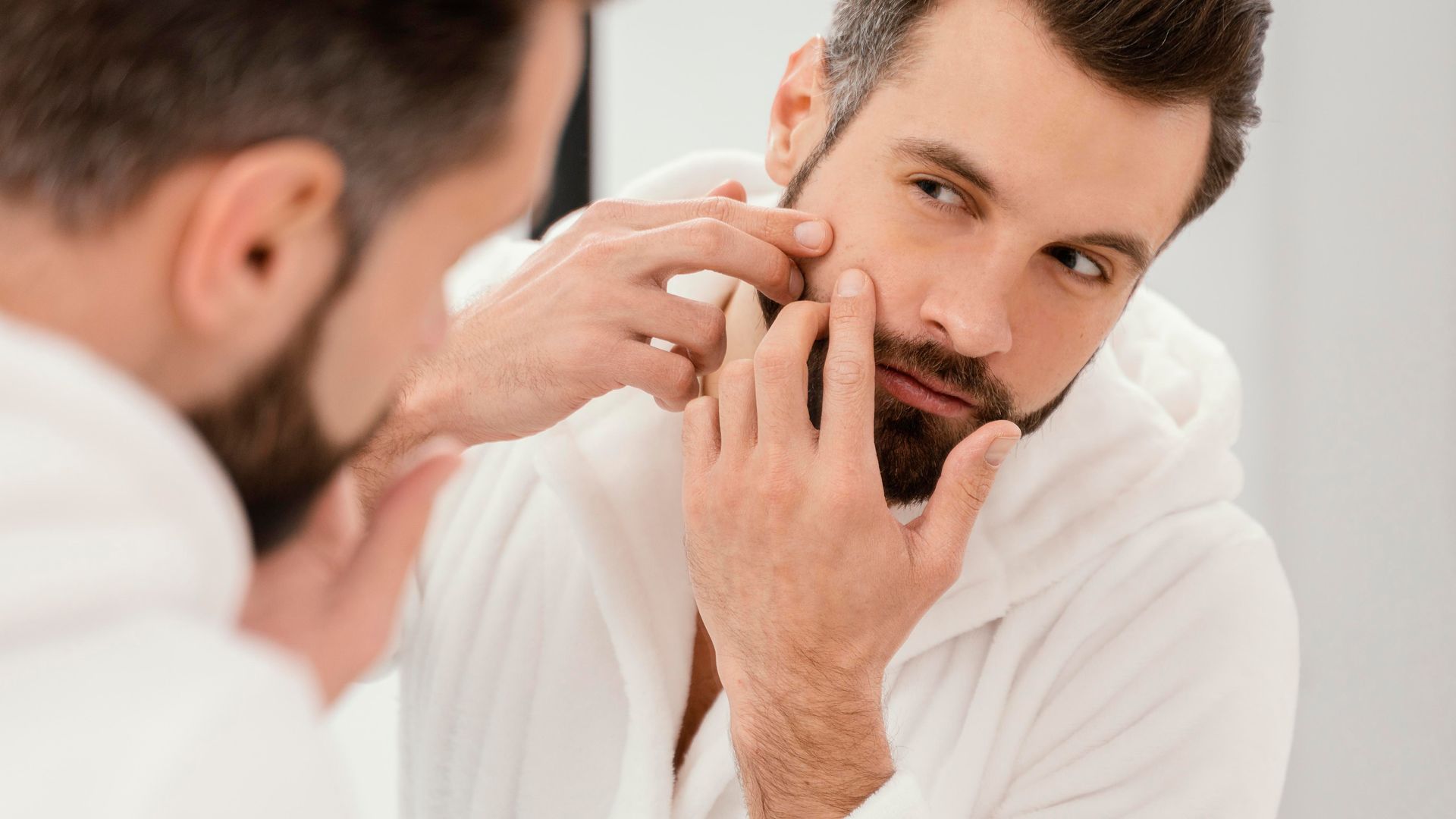 Man in white robe looking in mirror, examining face for blemishes.