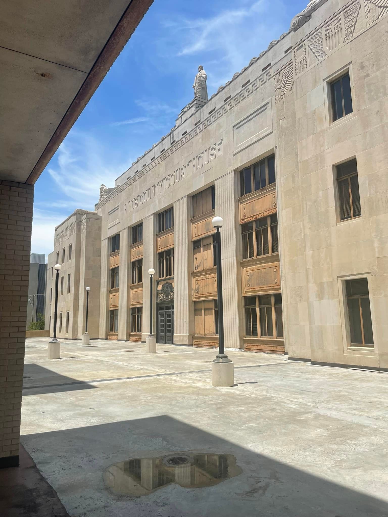 Exterior view of a weathered light-colored building with dark boarded windows, a sculpture on top, and a paved courtyard.