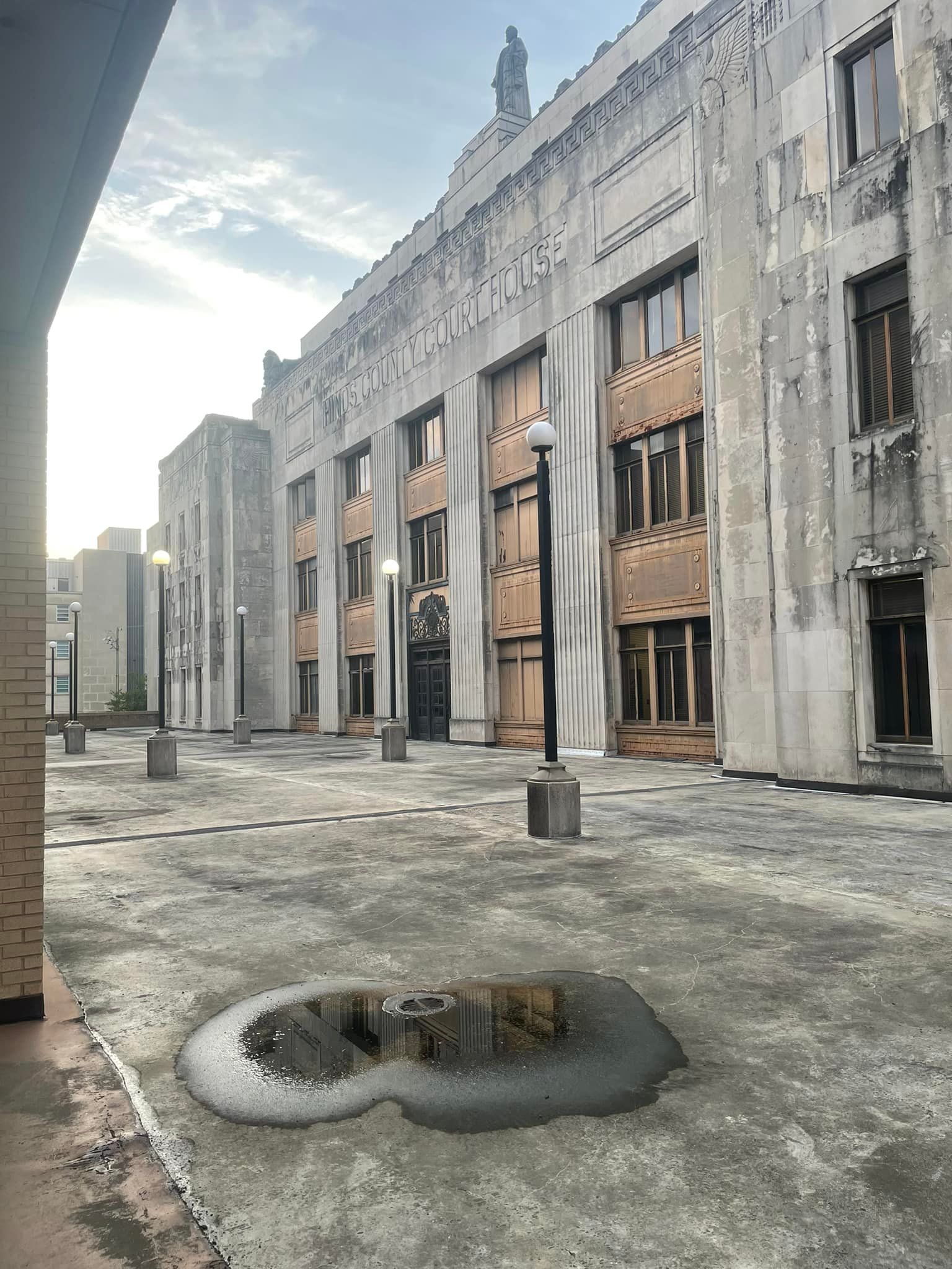 Exterior of a gray weathered building with multiple stories, light posts, and a puddle on the pavement.