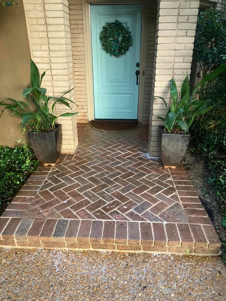 Brick pathway leading to a teal door with a wreath. Potted plants flank the entrance, set within a brick-pillared entryway.