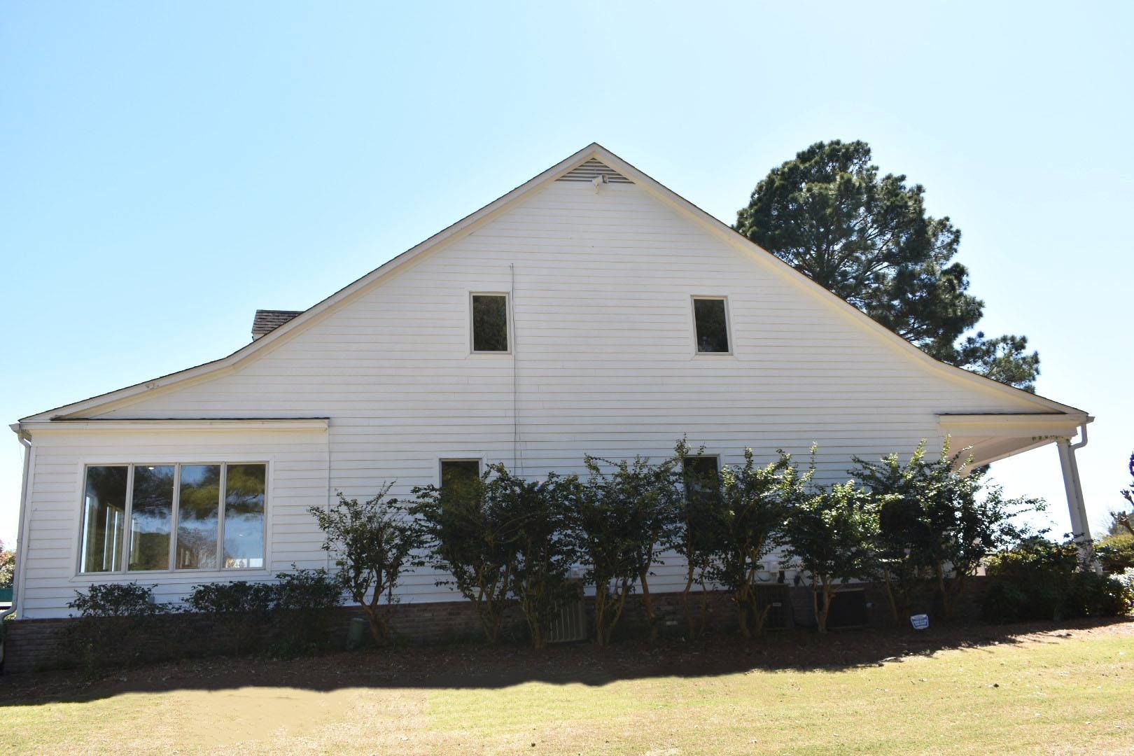 White house with a large gabled roof, side view. Porch and two small windows are visible.