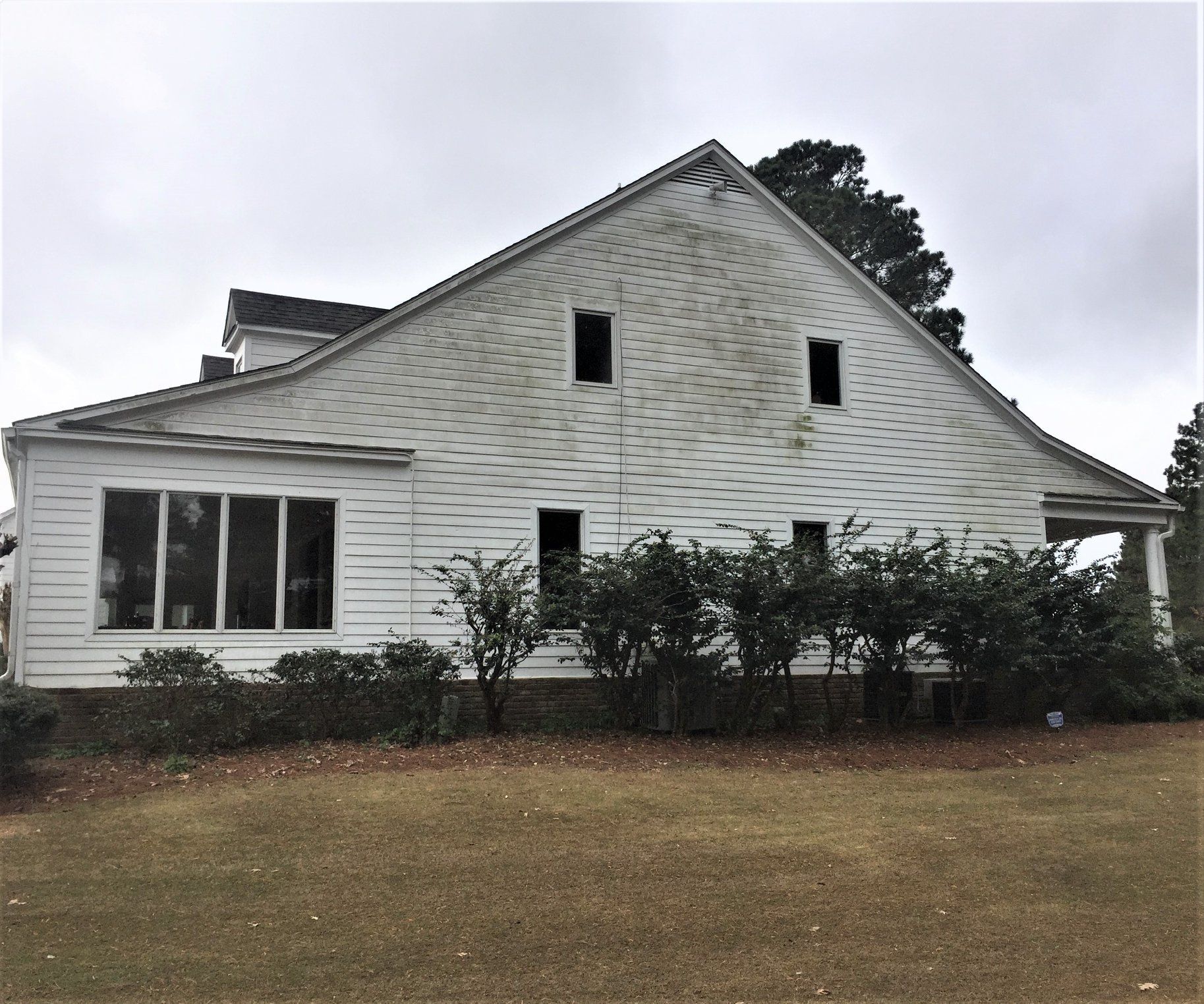 Side view of a weathered white two-story house with dark window openings, surrounded by bushes, on a cloudy day.