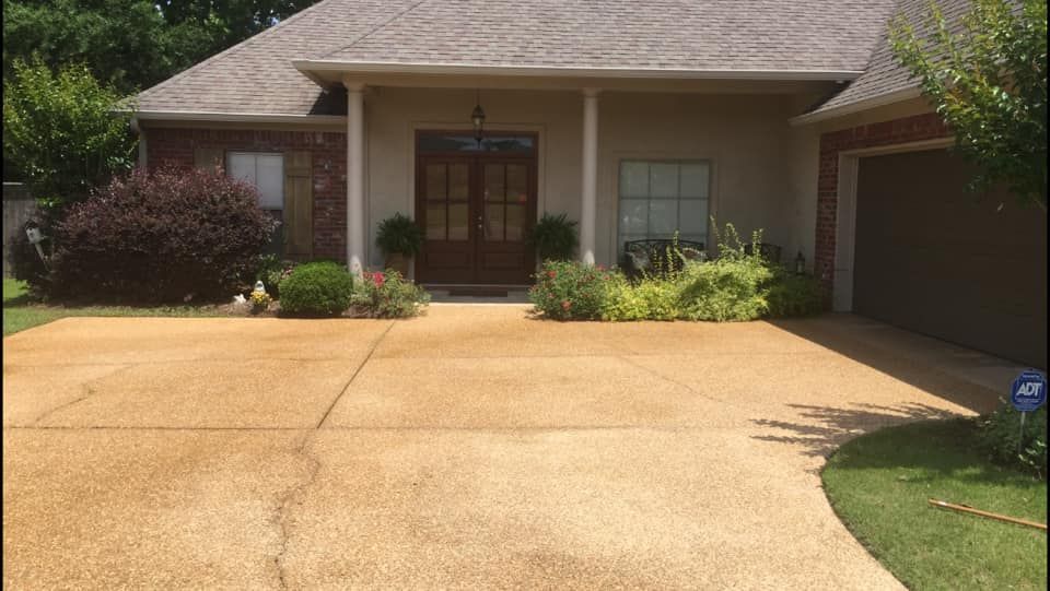 Tan concrete driveway leads to a beige house with a brown door. Landscaping surrounds the entrance.