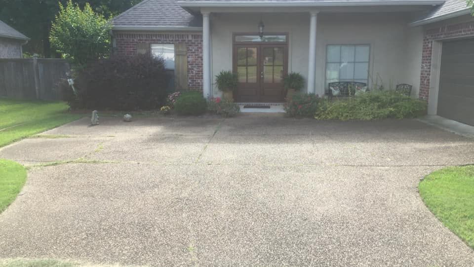 Driveway leading to a tan house with a brick accent and double doors; greenery surrounds.
