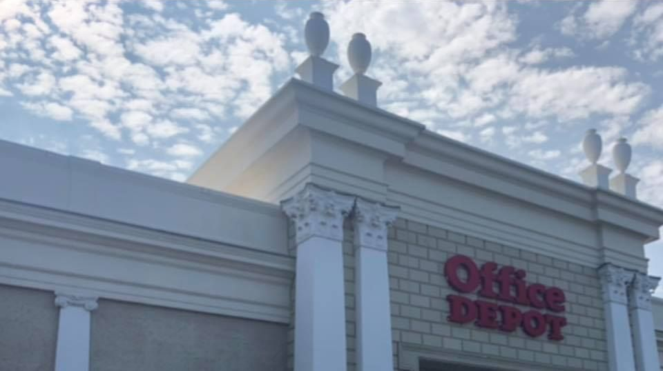 Office Depot storefront, white pillars and trim, red logo against a light-colored building, cloudy sky background.