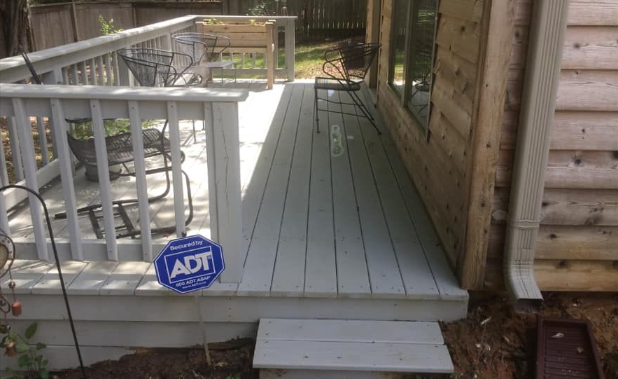 Wooden deck with white railing next to a house with a security system sign.