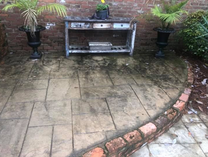 Patio with brick border and stained concrete slabs. A weathered table sits against a brick wall, flanked by potted plants.
