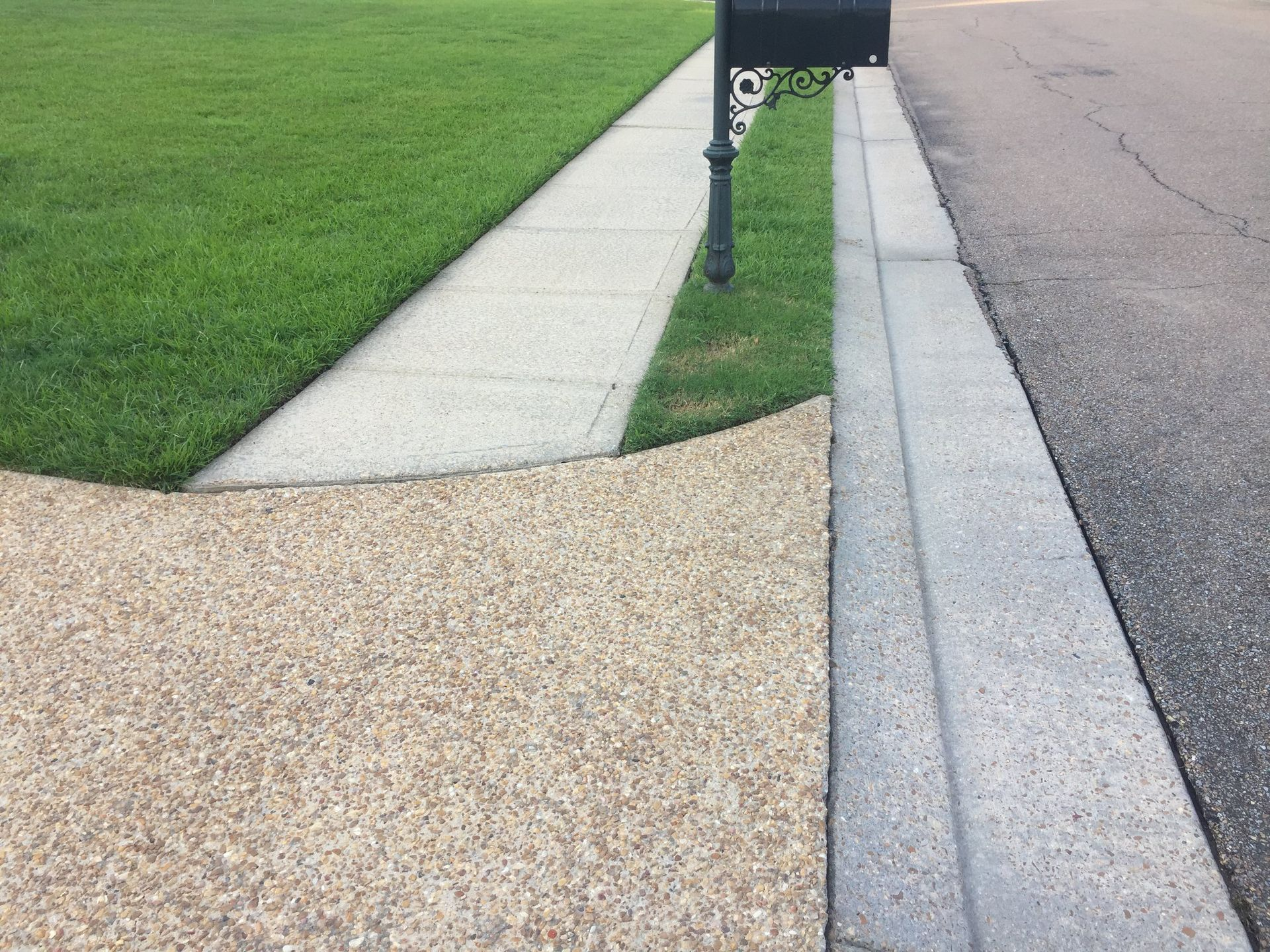 Sidewalk, grass, and street border a mailbox. The sidewalk is made of different textured concrete.