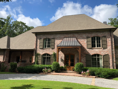 Brick house with green shutters, brown roof, and arched entryway under a metal awning. Lush green landscaping.