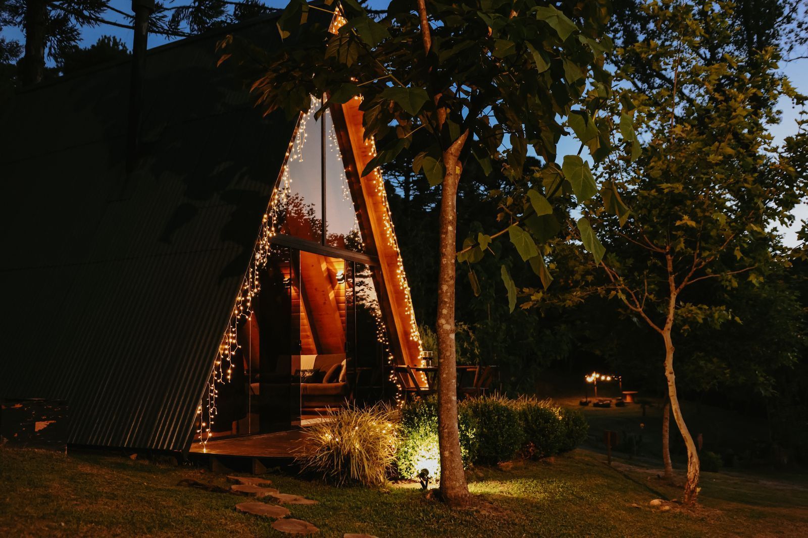 Uma cabana em forma de A, iluminada por luzes interiores aconchegantes, aninhada em uma paisagem arborizada ao entardecer.