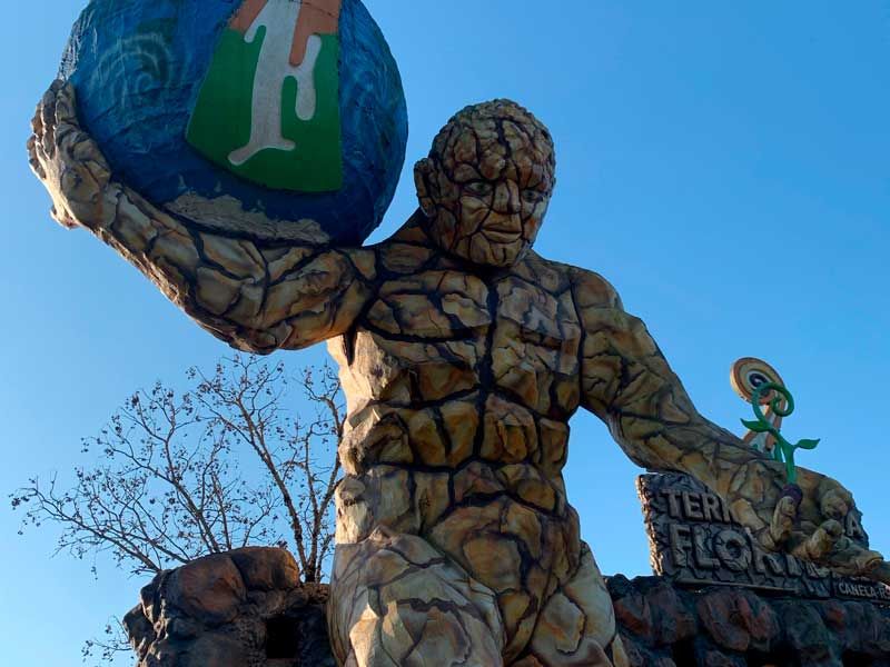 Estátua com textura de pedra representando uma figura musculosa segurando um globo terrestre contra um céu azul, com uma placa ao fundo.