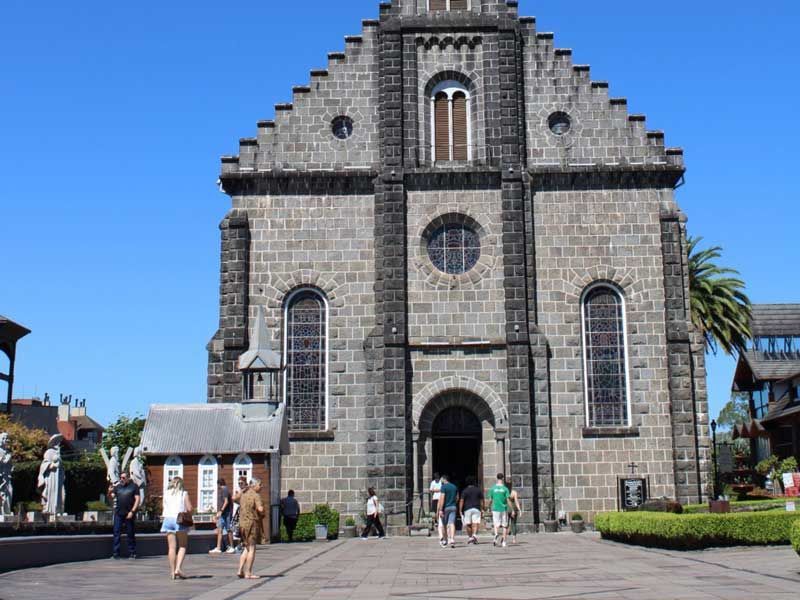 Uma igreja de pedra cinza, com telhado de duas águas escalonado e janelas em arco, ergue-se contra um céu azul, com pessoas caminhando em sua direção.