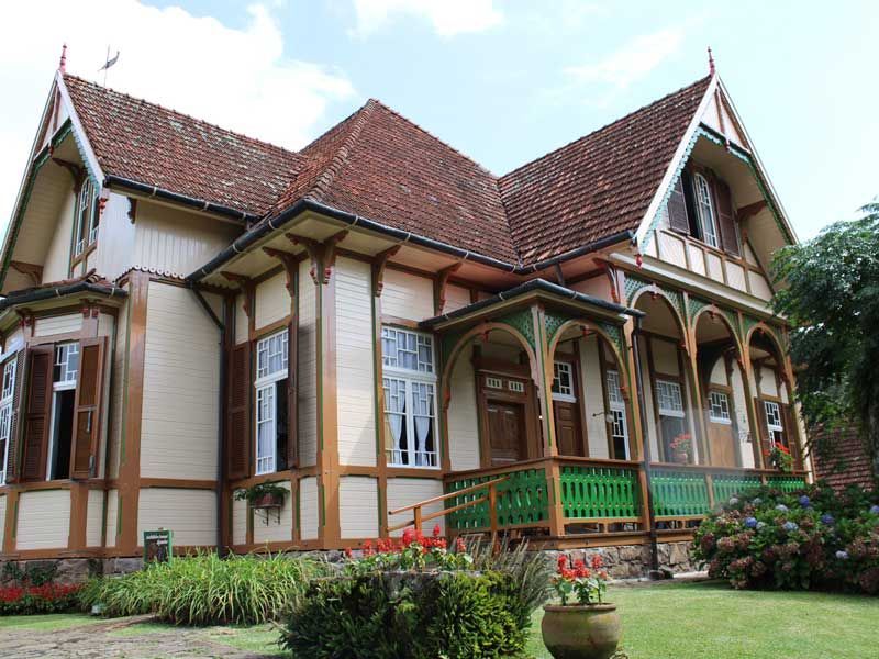 Uma casa histórica de madeira clara com detalhes em marrom, telhado de telhas vermelhas e varanda com grades verdes, situada em um jardim.