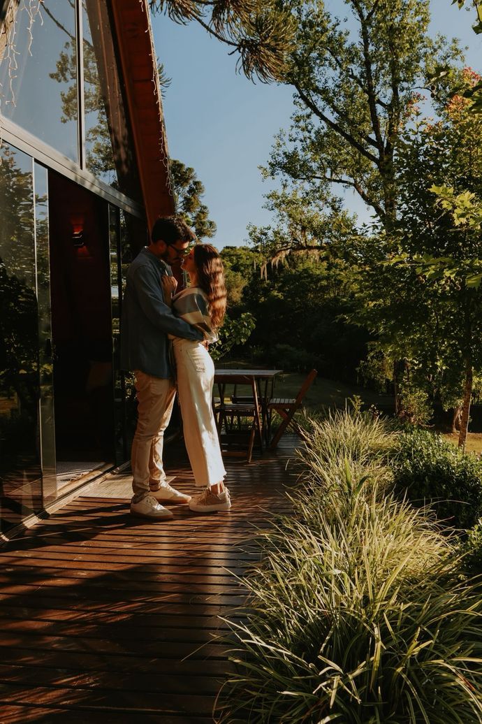 Um casal se abraça em um deck de madeira iluminado pelo sol, do lado de fora de uma cabana moderna em forma de A, cercada por árvores exuberantes.