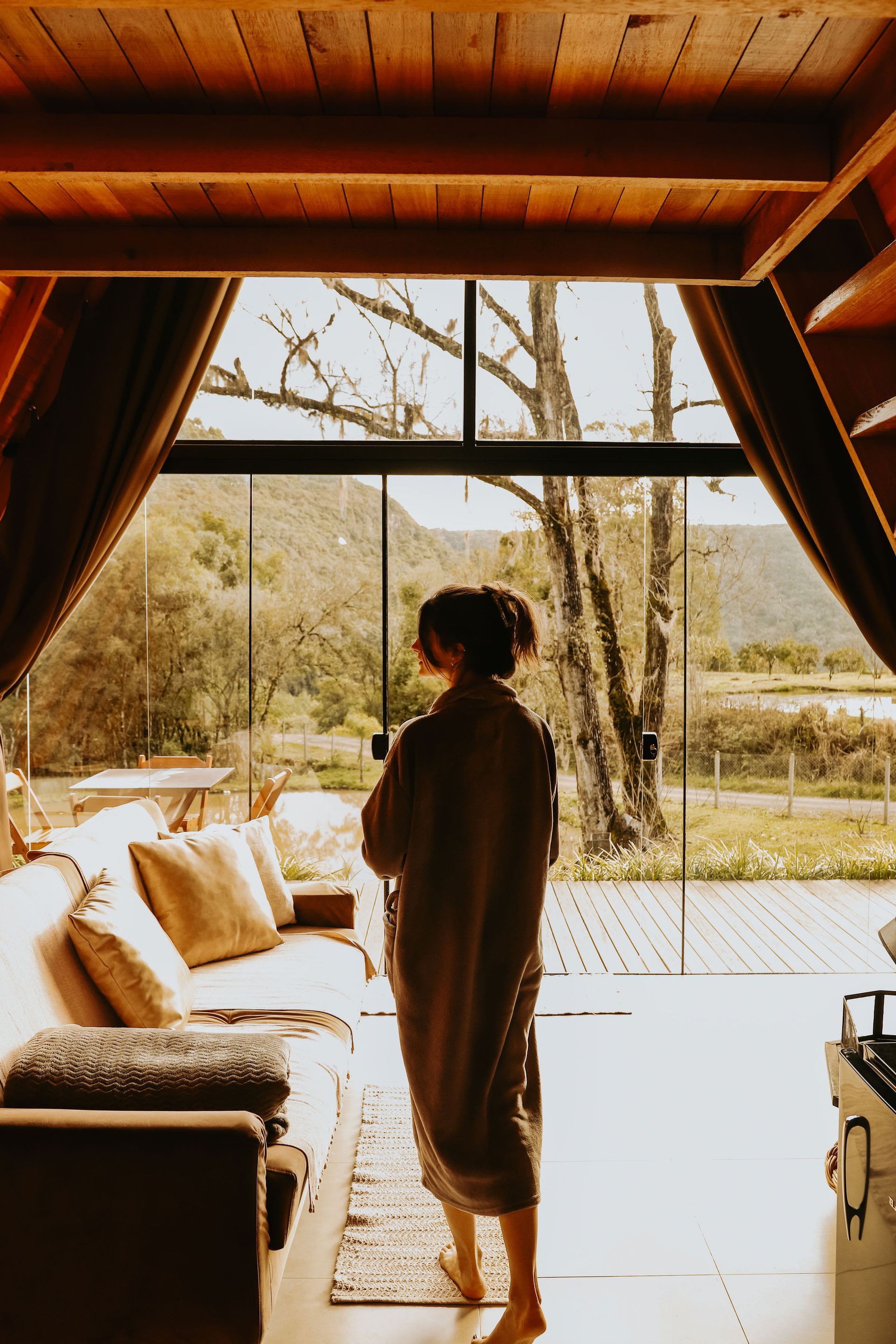 Uma pessoa vestida com uma túnica comprida está em uma cabana de madeira, olhando através de uma grande parede de vidro para uma paisagem externa deslumbrante.