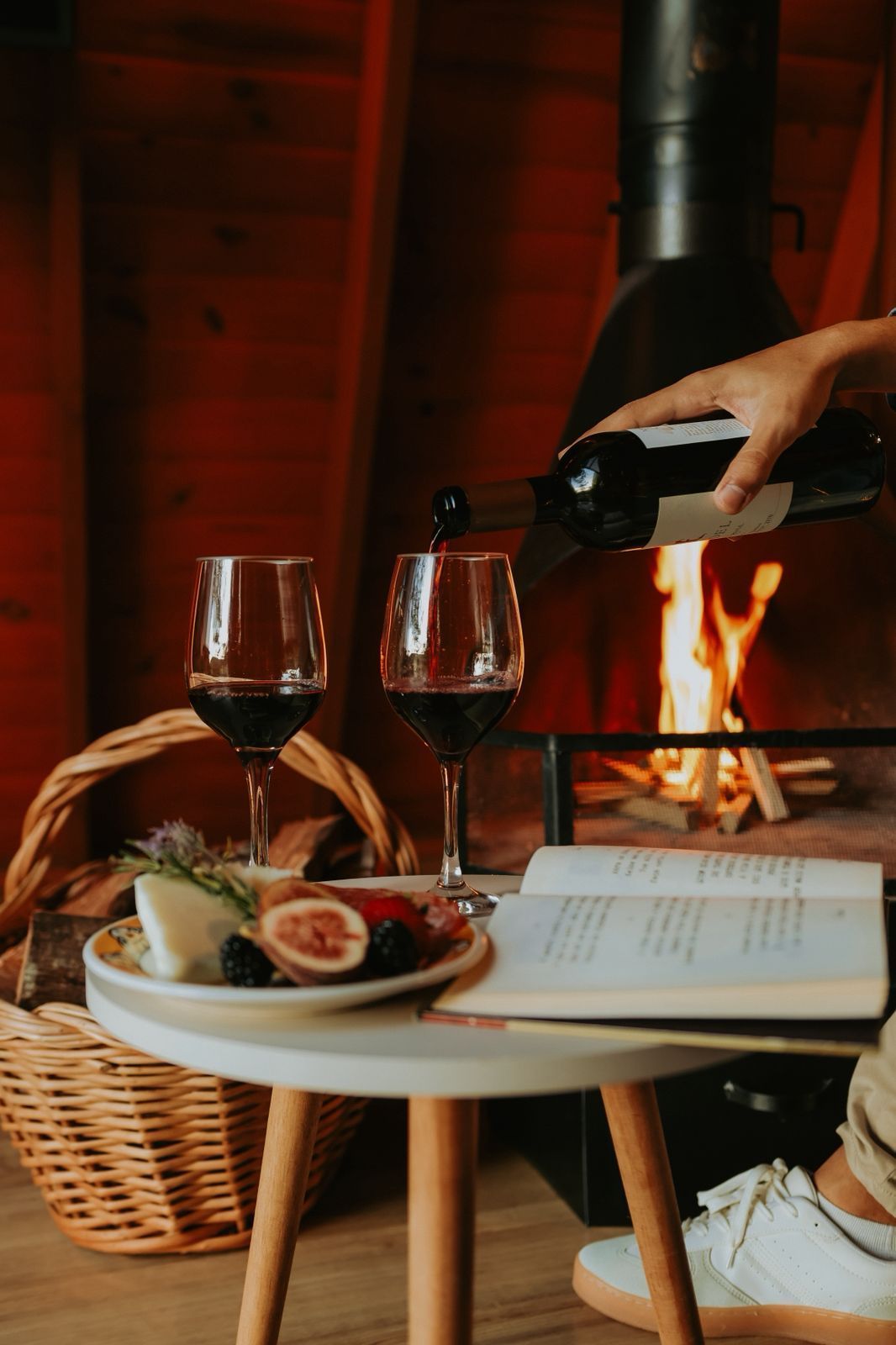 Uma pessoa serve vinho tinto em uma taça ao lado de um prato de frutas e um livro aberto, com uma lareira acesa ao fundo.