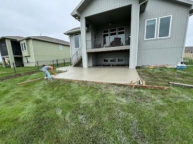 A person finishing a concrete patio next to a two-story house, on a grassy hill.