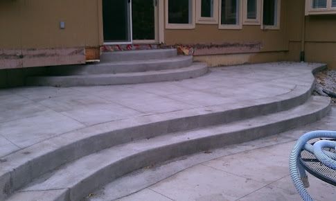 Concrete patio with curved steps leading up to a house with a sliding glass door and windows.
