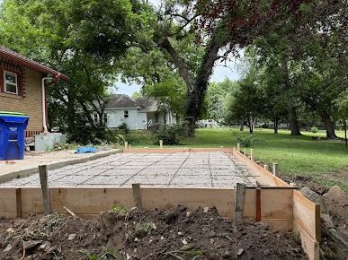 Wooden forms contain rebar grid for a concrete slab in a yard, with houses and trees in the background.
