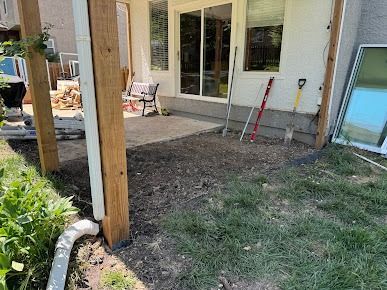 A patio with dirt and grass in front of a house. Lumber supports a roof. Tools lean against the wall.
