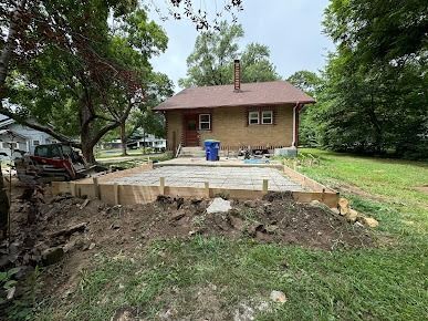 Concrete foundation with wooden frame in front of a brick house. Green grass and trees surround it.