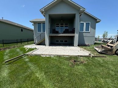 Back of gray house with a partially removed patio, construction equipment nearby, and green grass.