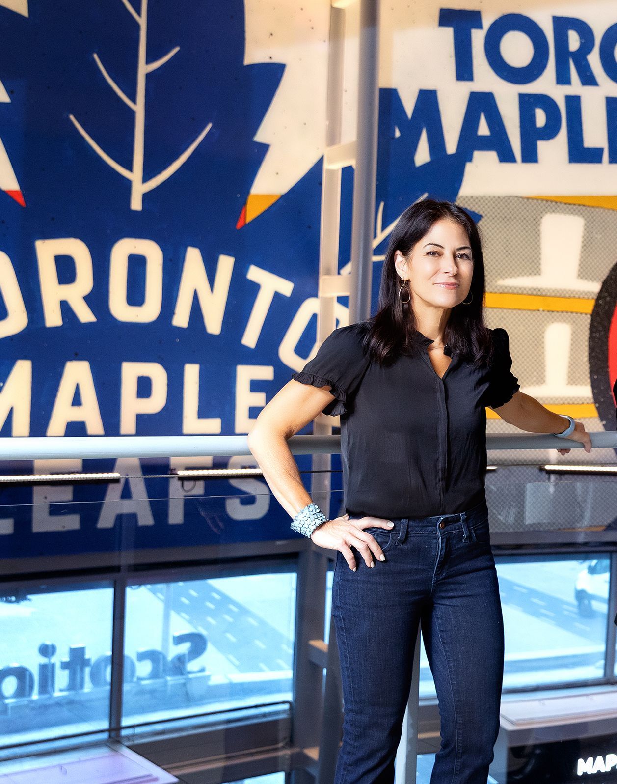 Woman stands near a Toronto Maple Leafs logo. She wears dark jeans, black top, and has a bracelet on her left wrist.