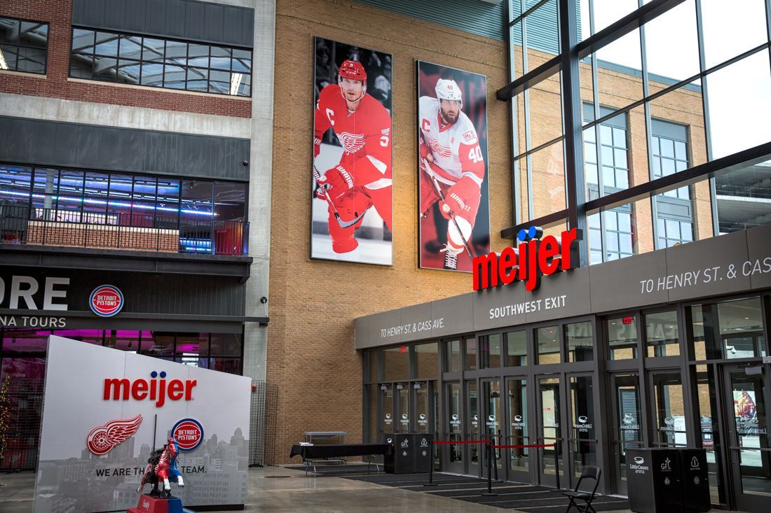 Meijer entrance at a building with Red Wings player banners. Brick wall and glass windows visible.