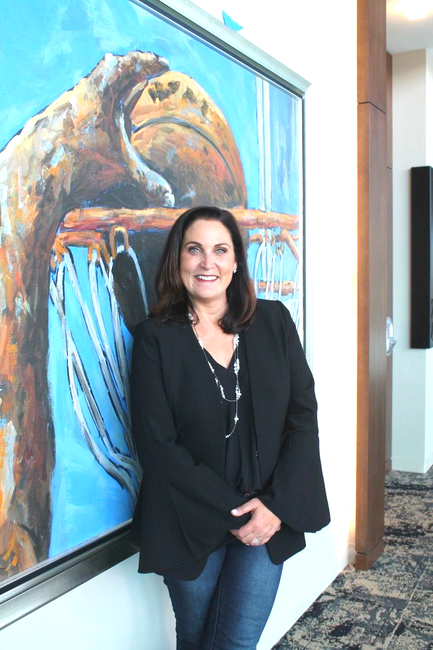 Woman in black blazer and jeans stands beside an abstract painting with blue and brown hues.