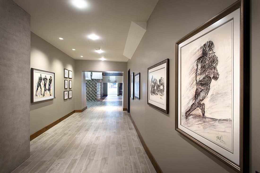 Hallway with framed artwork on gray walls, light-colored wood floor, and recessed ceiling lights.