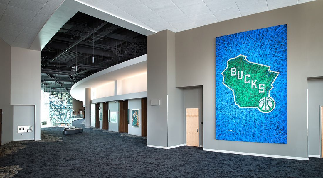 Lobby with Milwaukee Bucks logo art on a wall; dark carpet, gray columns, and doors.