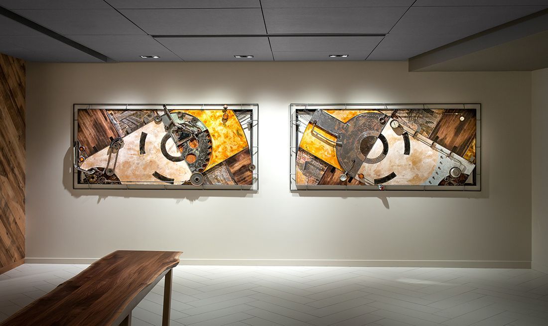 Two framed abstract artworks hang on a white wall in a gallery. A wooden bench sits below.
