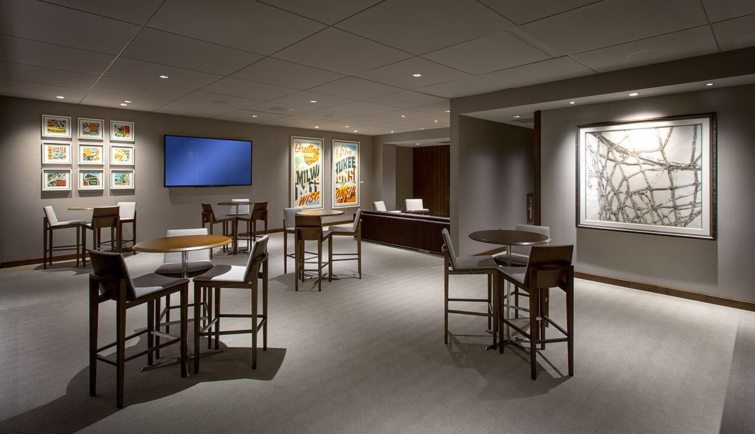 A dimly lit bar area with high tables and stools. Artwork and a blue screen are visible on the walls.