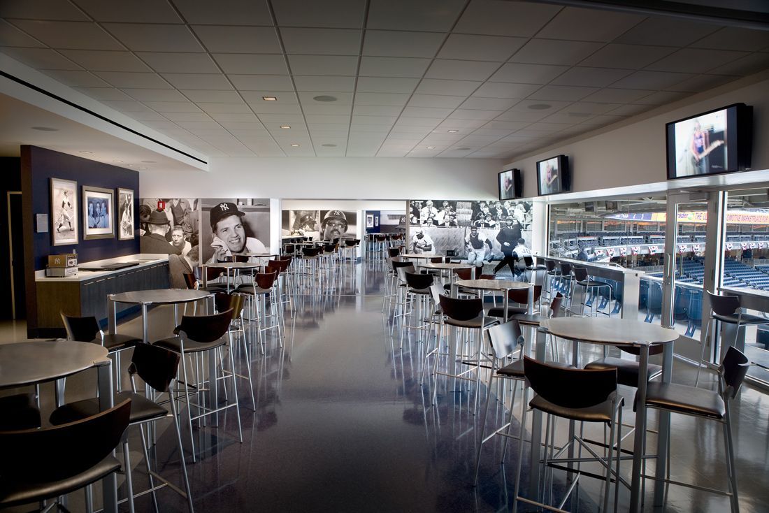 A stadium lounge with tables and chairs. Large windows overlook a sports field. TVs and artwork adorn the walls.