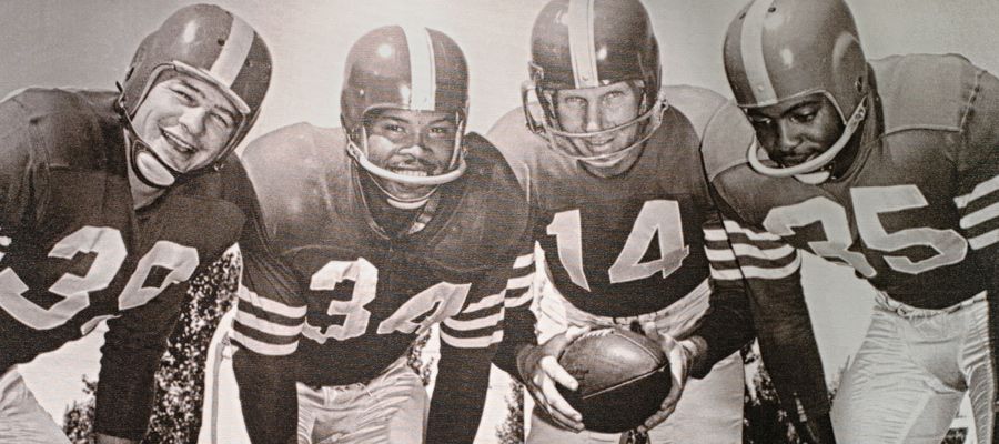 Four football players in helmets and jerseys, holding a ball.