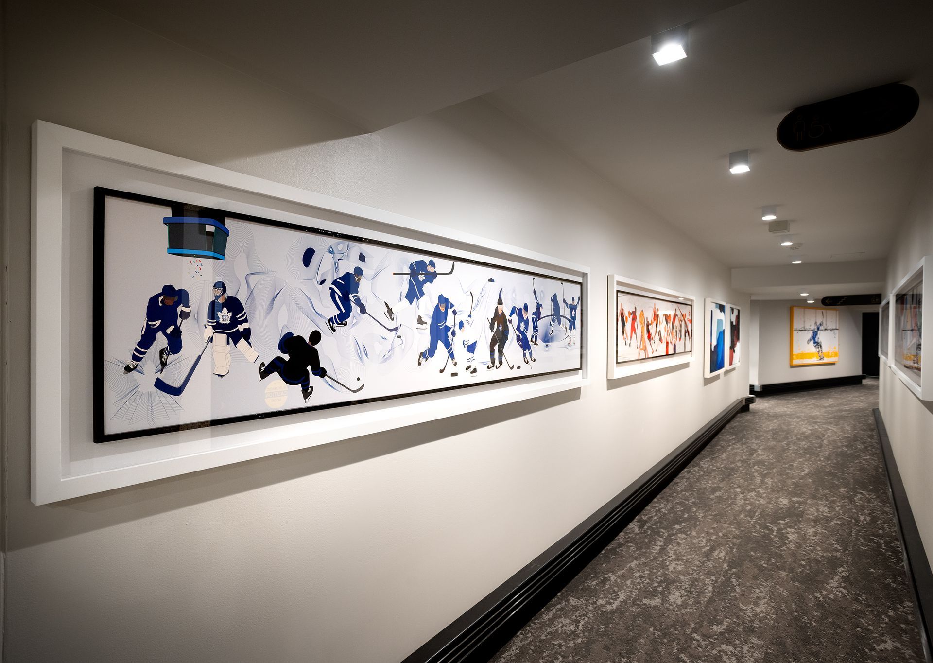 Hallway with several framed hockey-themed art pieces. White walls, black trim, and dark patterned carpet.