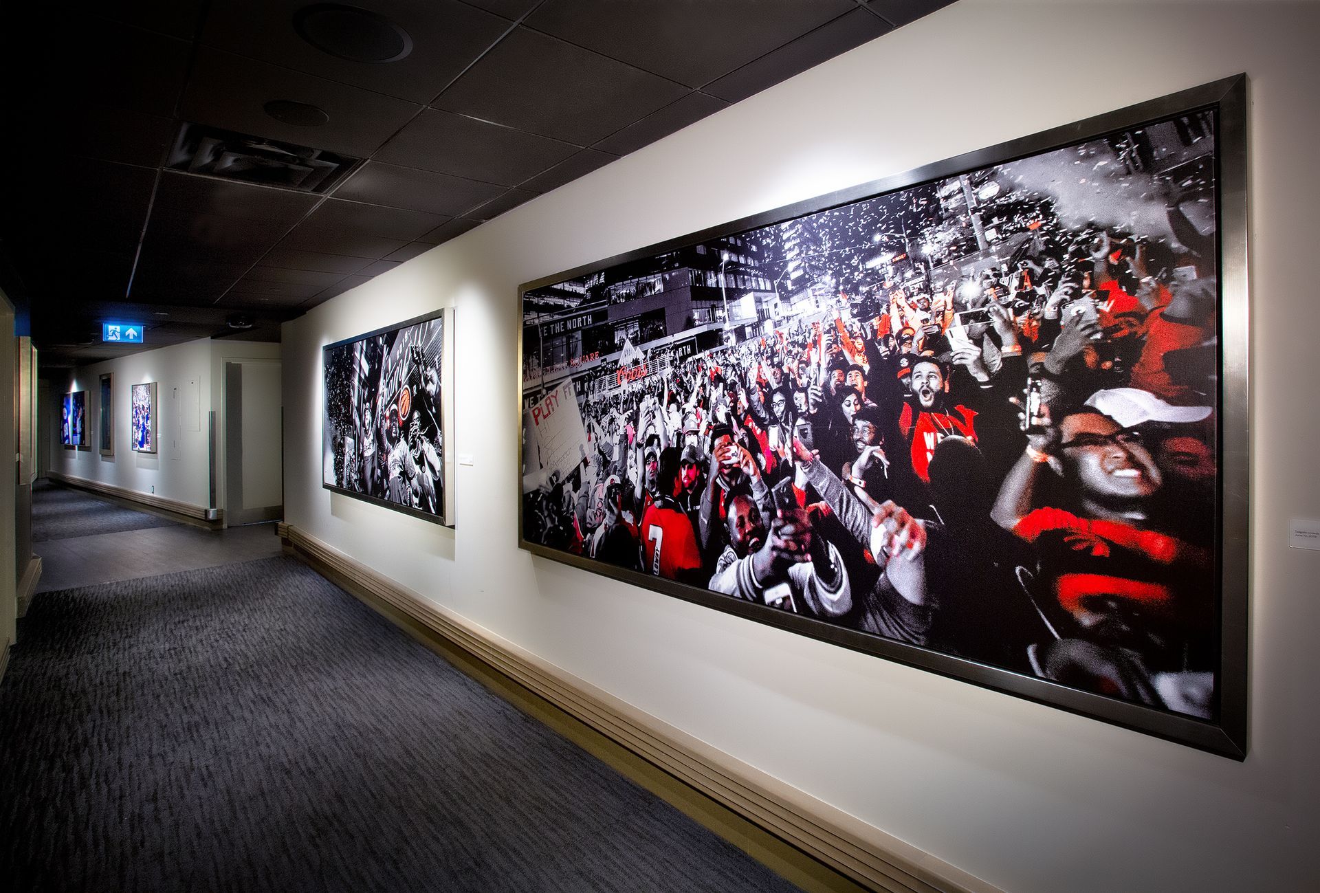 Hallway with framed black-and-white photos of a crowd, some in red, illuminated by overhead lights.