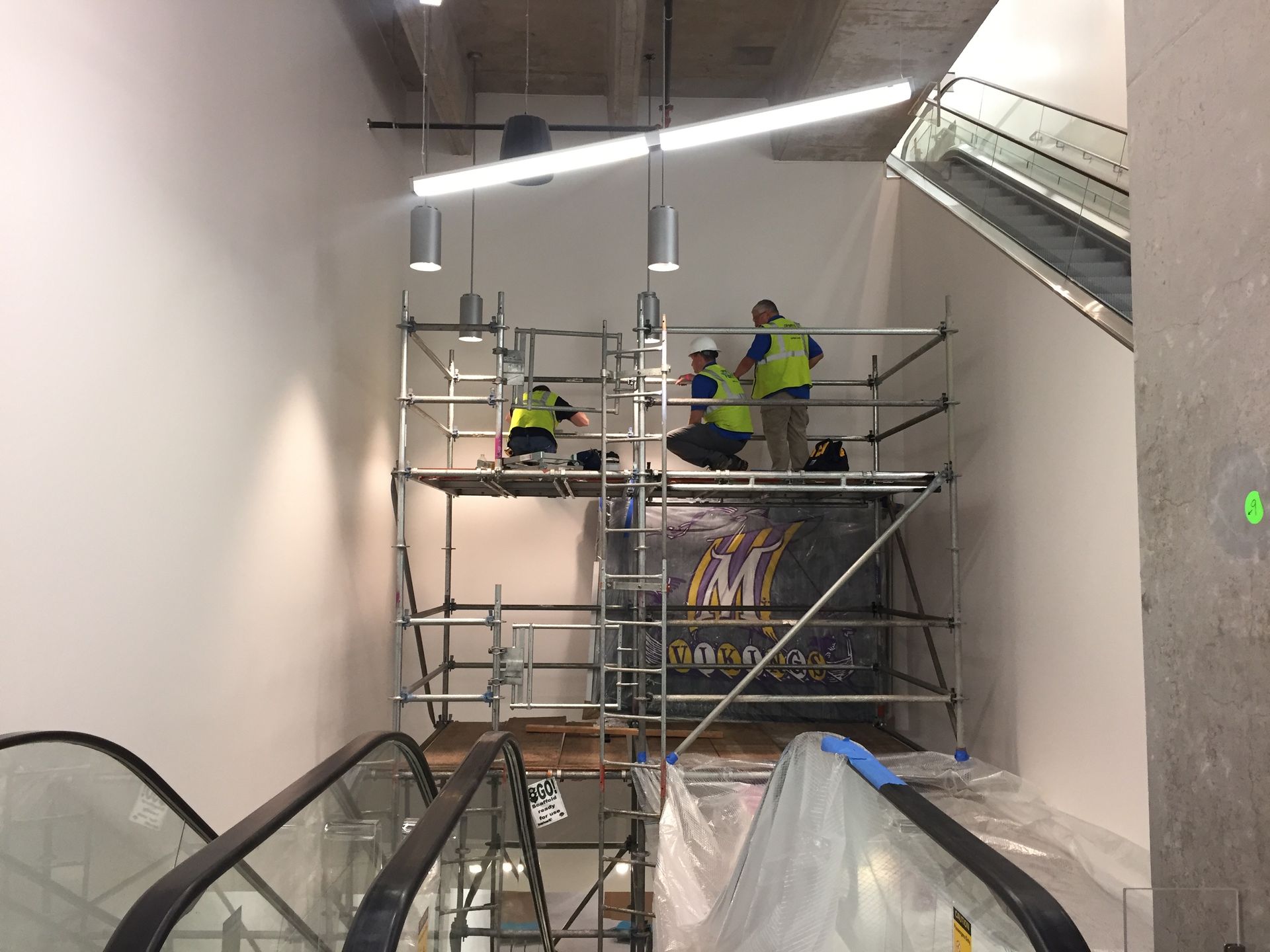 Workers on scaffolding near an escalator. They are wearing safety vests and working on lighting.