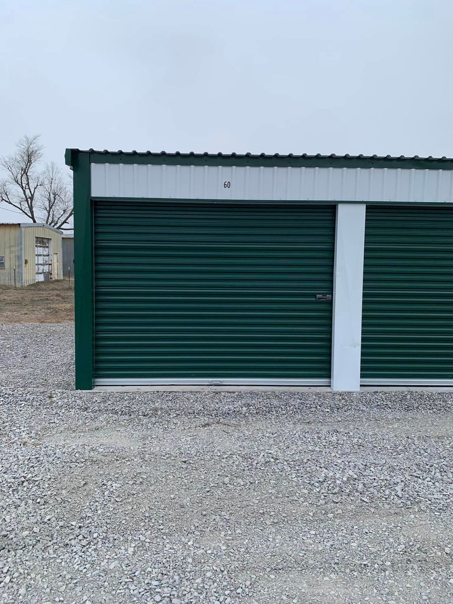 A view of green metal storage unit doors in a gravel lot under a cloudy sky.