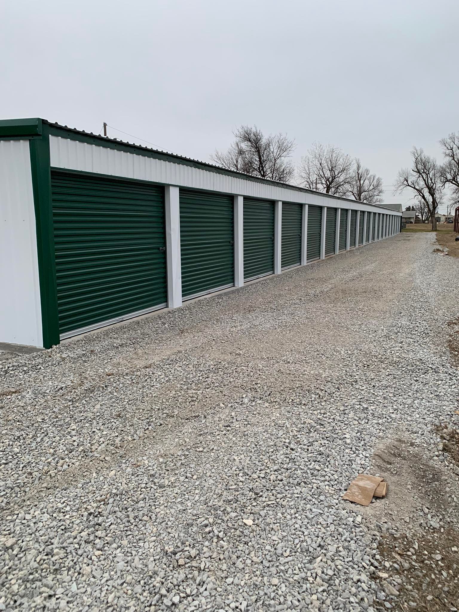 A row of self-storage units with dark green roll-up doors and white walls, situated on a gravel lot under an overcast sky.