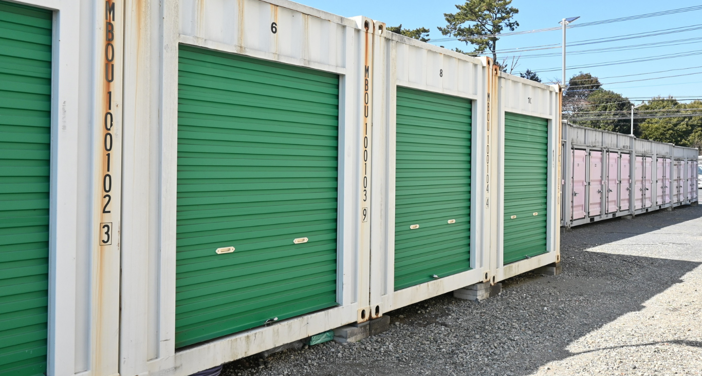 A row of white shipping container storage units with green roll-up doors outdoors on a gravel lot under a clear blue sky.