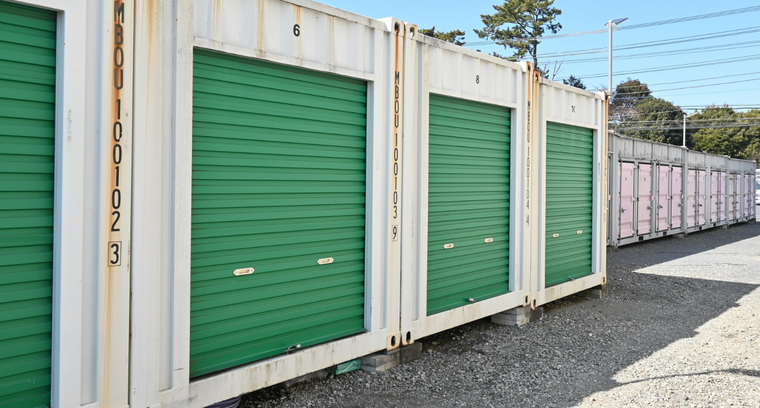 A row of white shipping container storage units with green roll-up doors outdoors on a gravel lot under a clear blue sky.