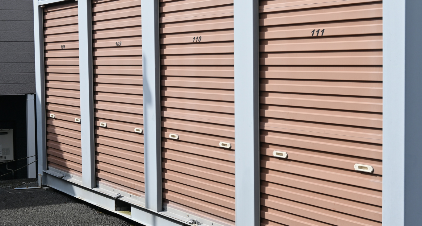 A row of tan, corrugated metal roll-up doors for self-storage units with white trim, located outdoors.