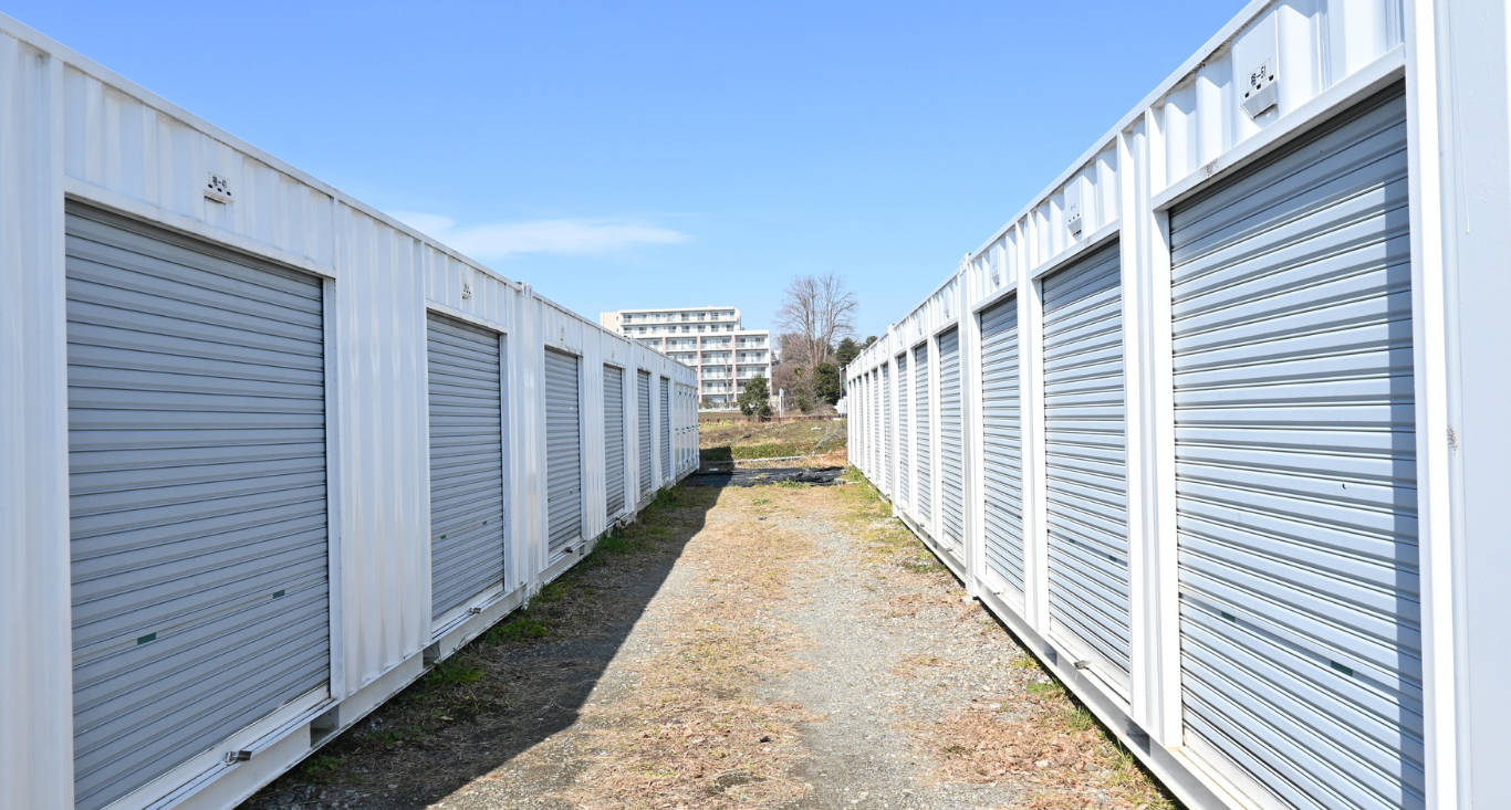 Two rows of white storage container units with grey roll-up doors face each other across a dirt path under a clear sky.