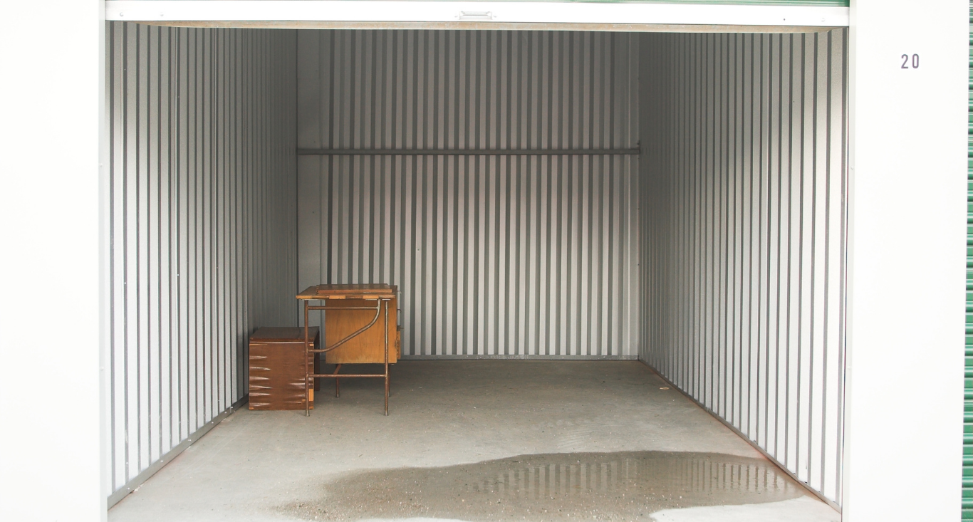 An empty storage unit with white corrugated walls, containing a small wooden desk and a chair.
