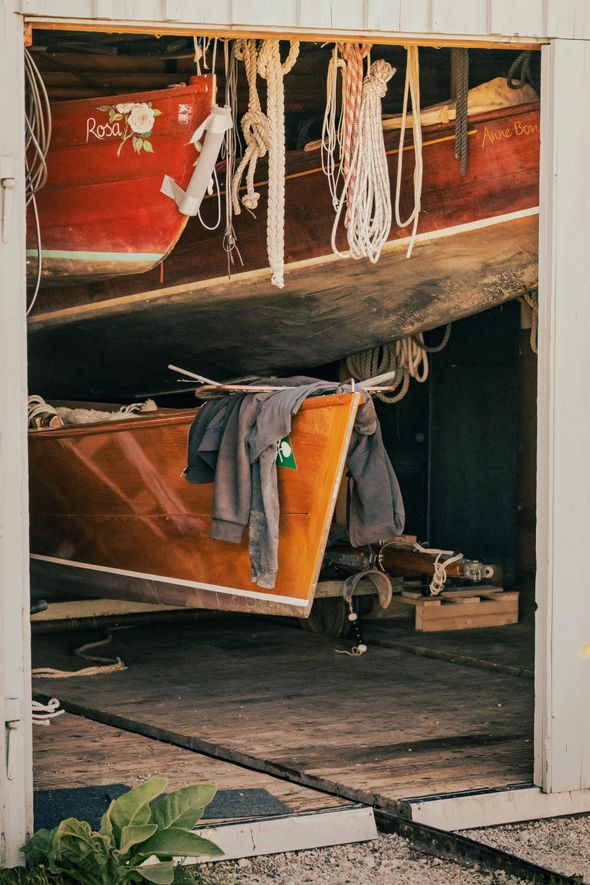 A boat shed contains a red boat suspended above a wooden boat with a grey garment draped over its bow.