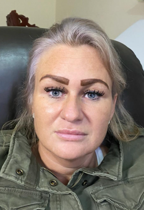 Woman with blonde hair, gray roots, brown eyebrows, and wearing a green jacket, looking forward.