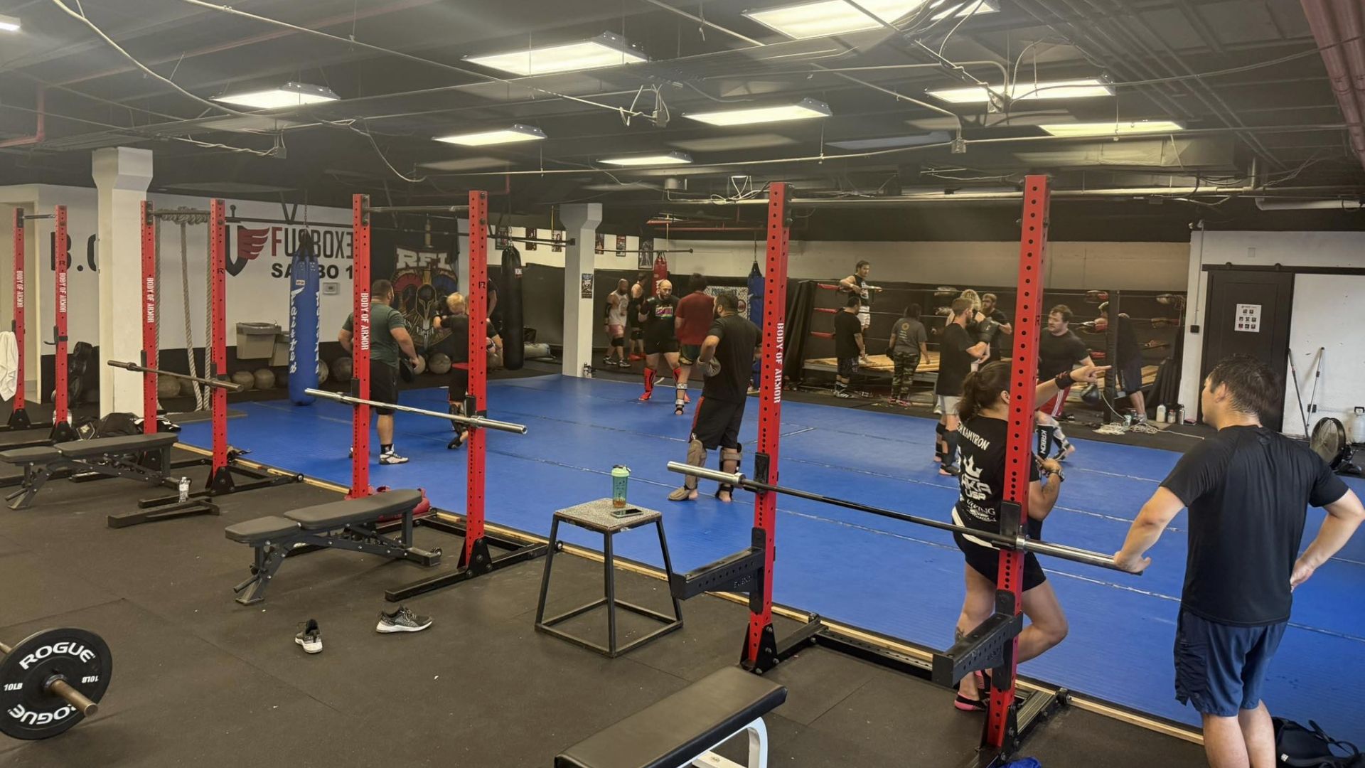 People train in an indoor gym with red squat racks, a blue floor, and exercise equipment.