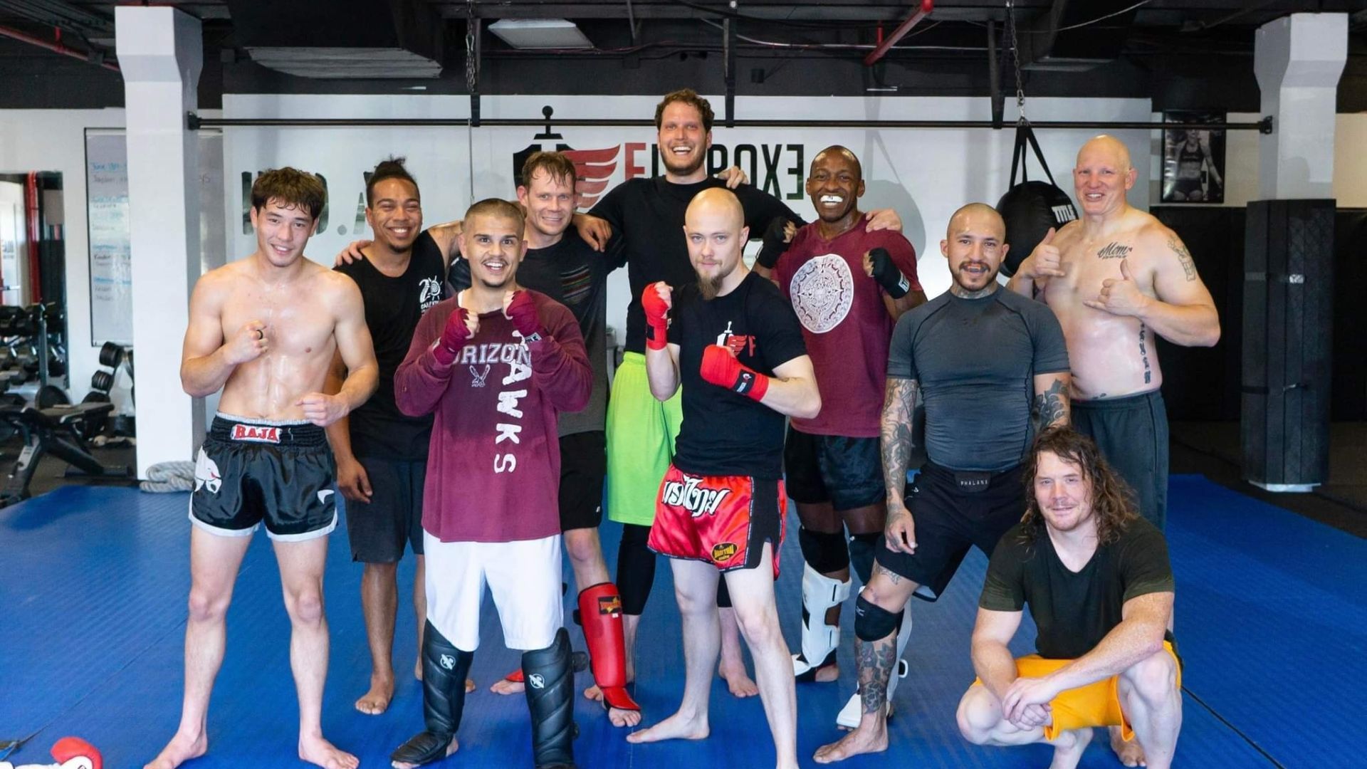 Group of fighters posing for a photo in a gym, some wearing training gear, standing on a blue mat in front of equipment.