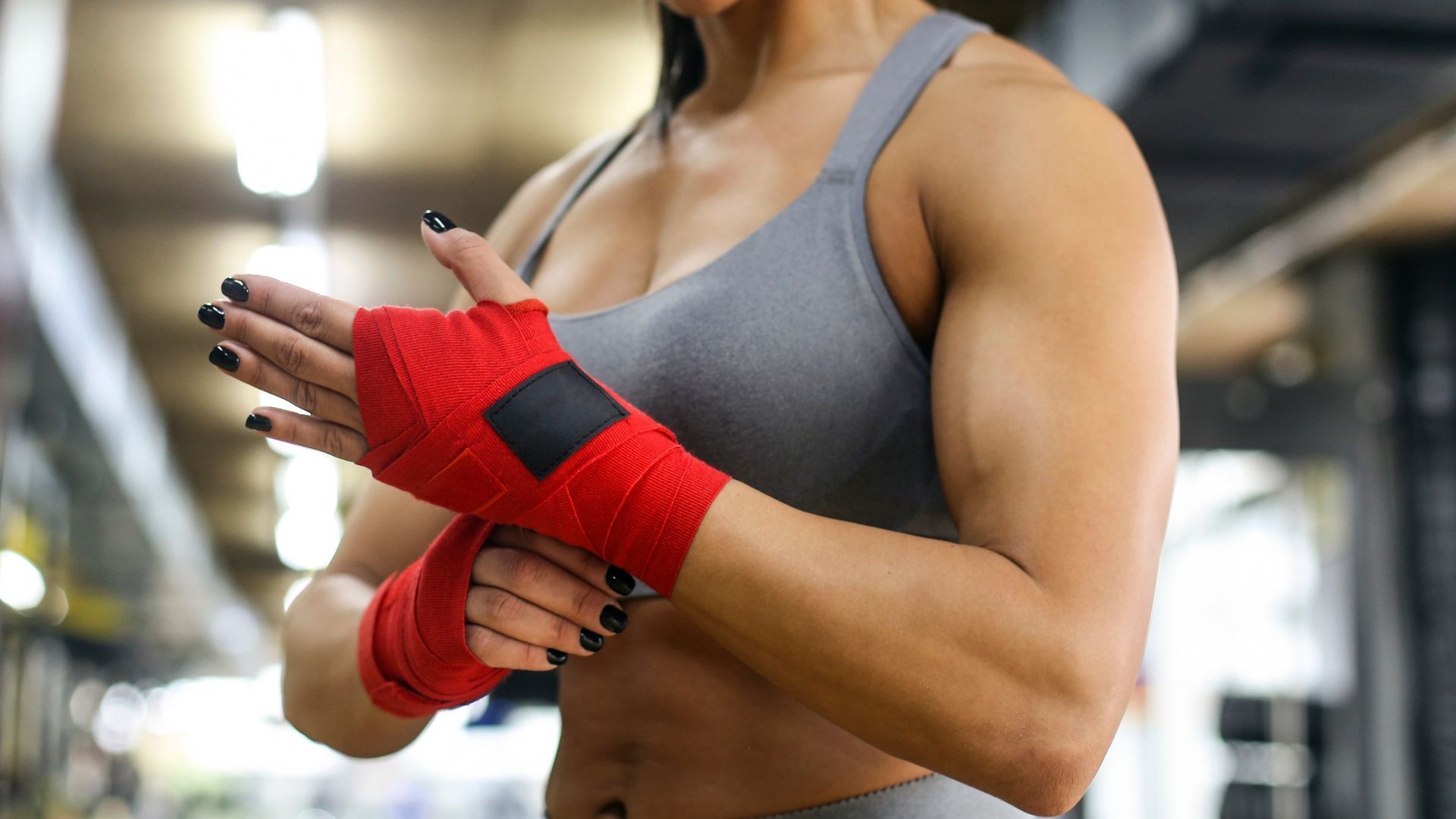 An athlete in a gray sports bra wraps their hands with red boxing bandages inside a gym.